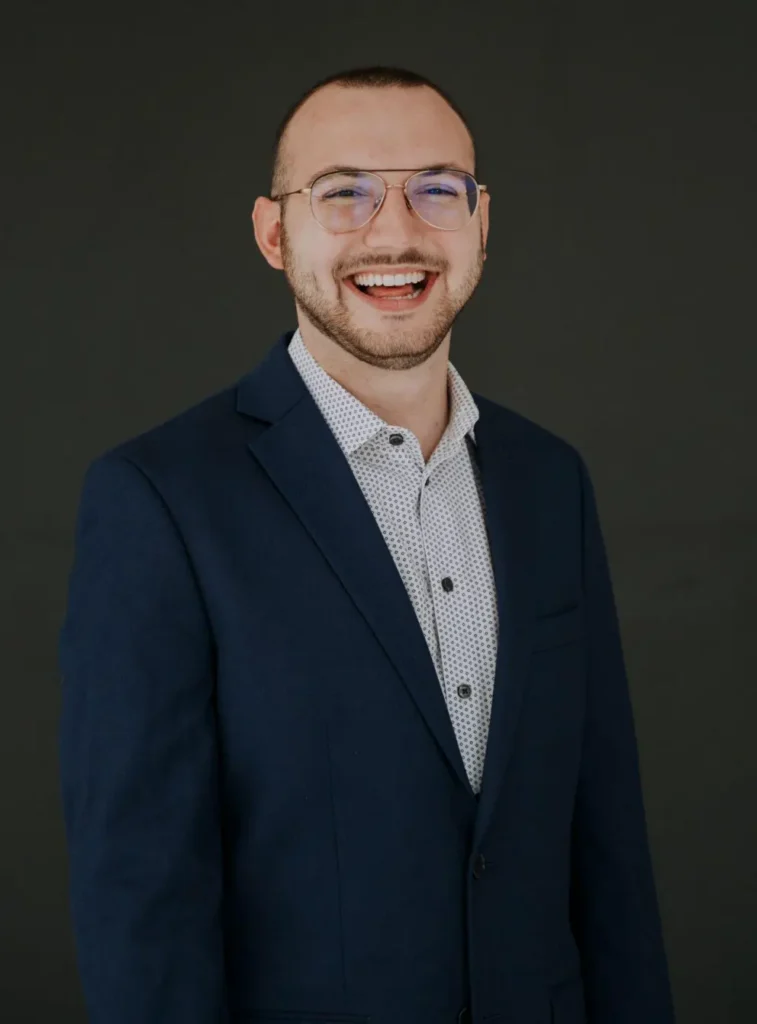 Smiling man in a navy blazer and patterned shirt facing the camera against a dark background.